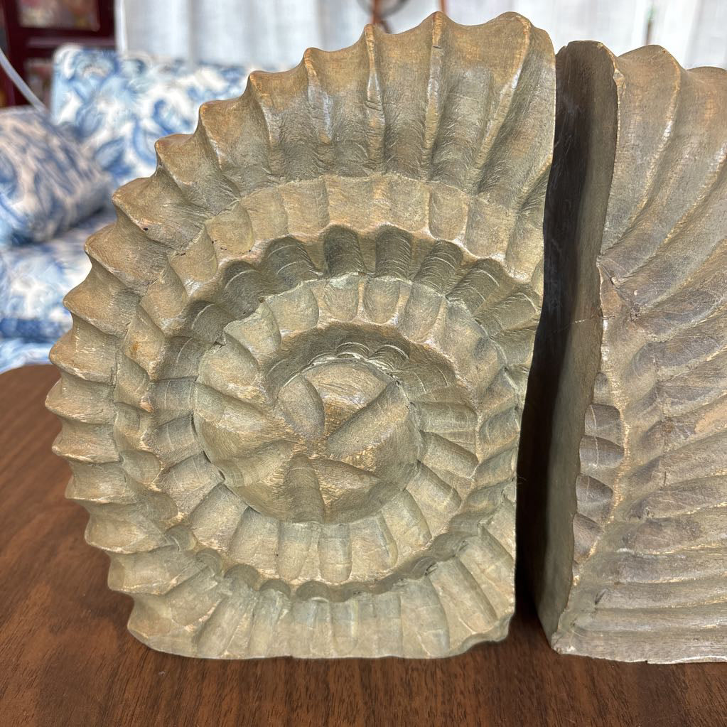 A pair of carved stone bookends shaped like coiled ammonite or seashell structures, displayed on a wooden table with a blurred living room background.