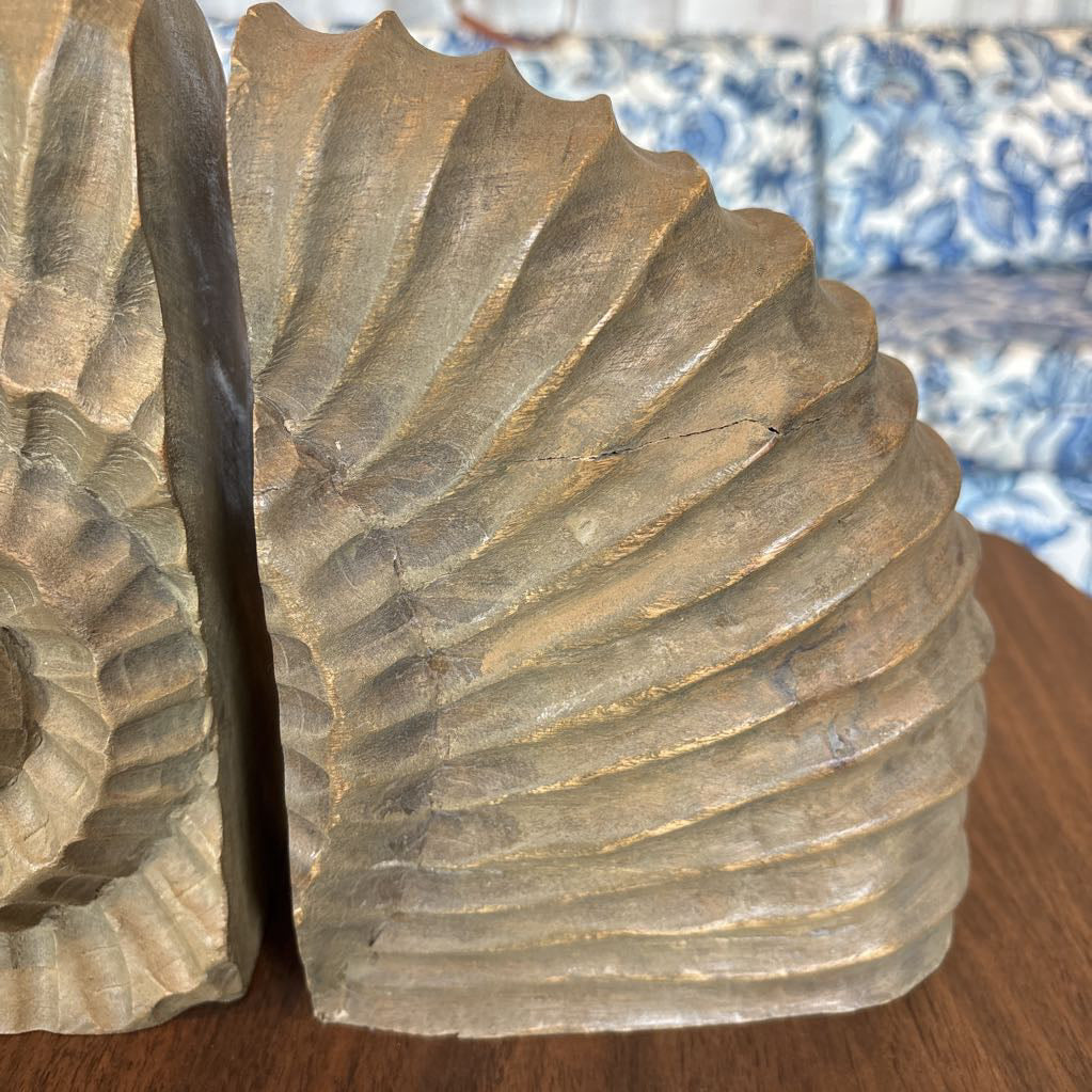 A close-up of a carved wooden bookend shaped like a seashell, showing detailed grooves and a natural wood grain finish, sitting on a wooden table with a blue and white patterned sofa in the background.
