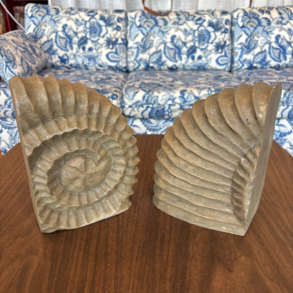 A close-up of a carved wooden bookend shaped like a seashell, showing detailed grooves and a natural wood grain finish, sitting on a wooden table with a blue and white patterned sofa in the background.