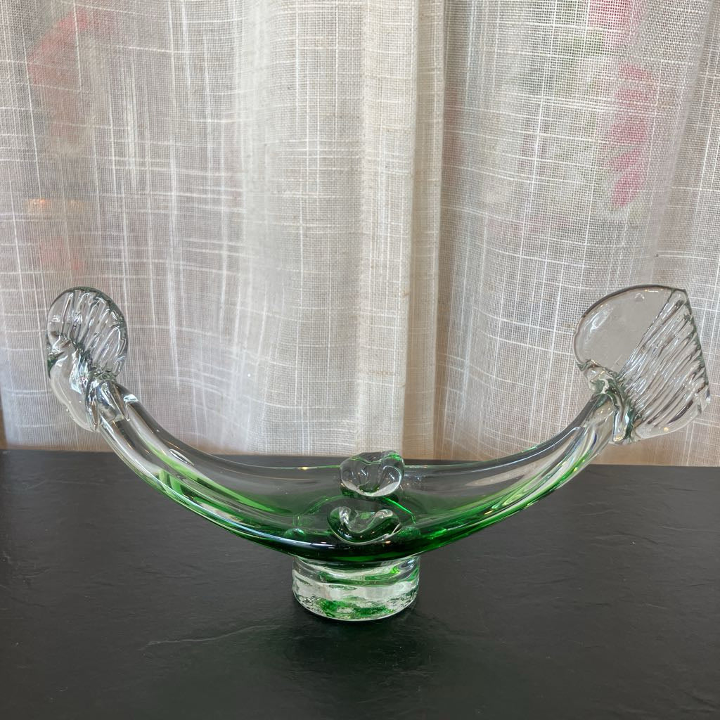 A vintage green-tinted clear glass boat-shaped bowl with ribbed ends, displayed on a dark surface in front of a beige curtain backdrop.