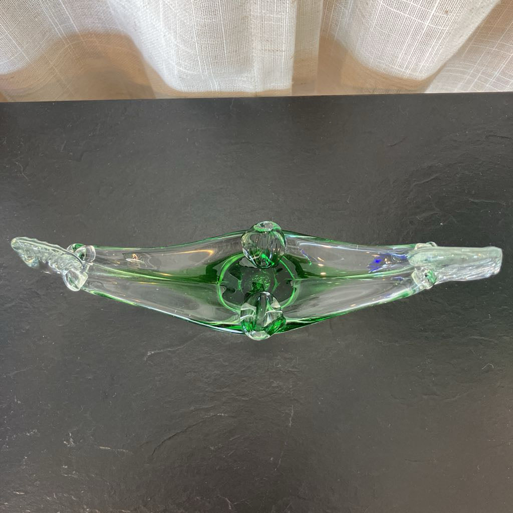 A green-tinted clear glass decorative object with a sculptural, winged or leaf-like form, photographed on a dark tabletop with a light curtain in the background.