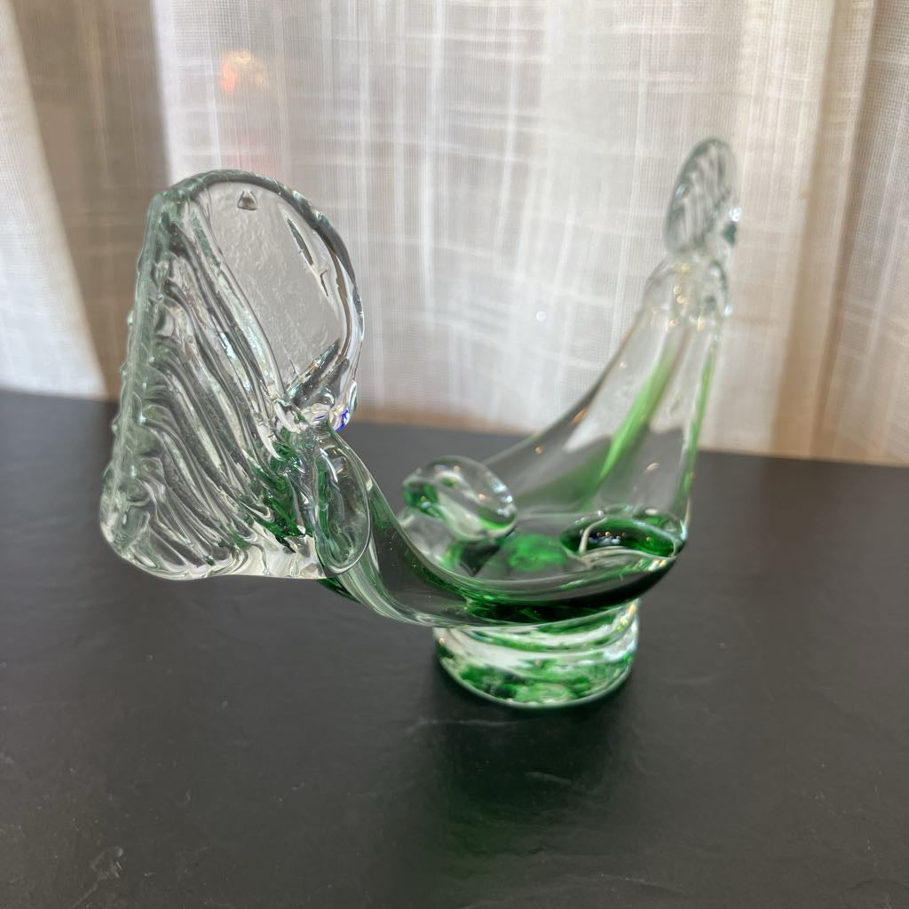 A clear glass bird figurine with vibrant green accents, displayed on a dark surface in front of a light-colored curtain background.