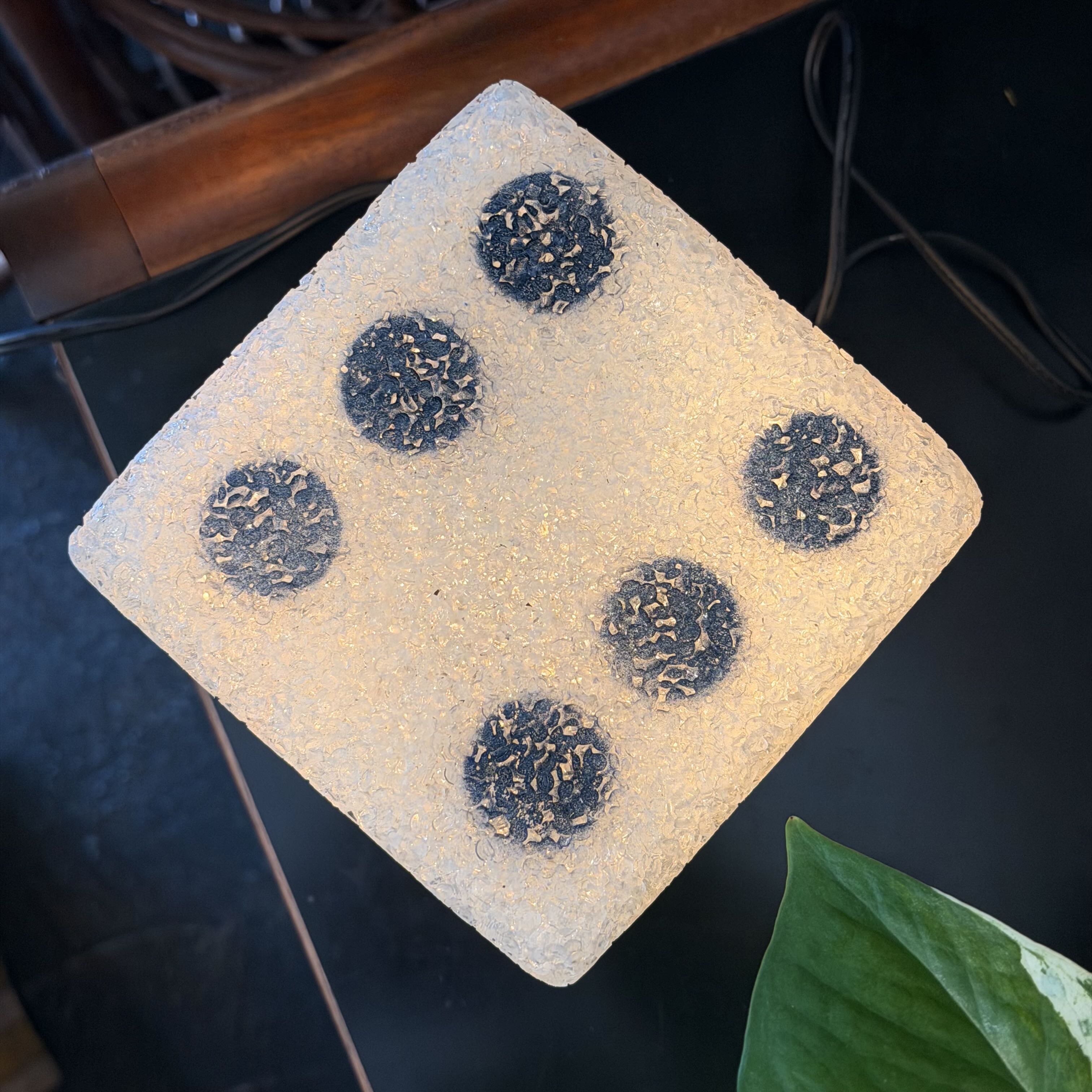 A square, diamond-angled decorative plaque or coaster made of textured beige stone or resin, featuring five dark gray circular spots with small beige inclusions, shown on a dark tabletop.