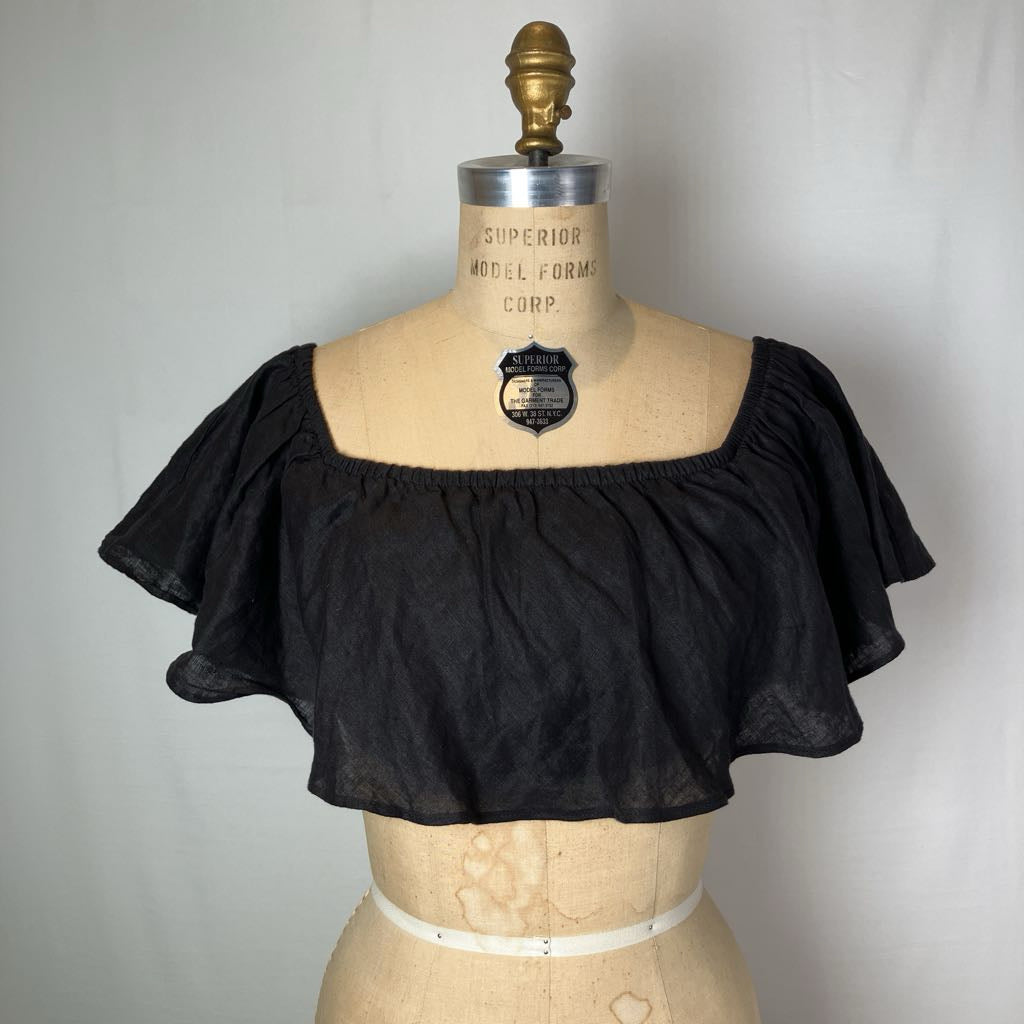 A black off-the-shoulder ruffled crop top displayed on a vintage wooden dress form mannequin against a light gray fabric backdrop.