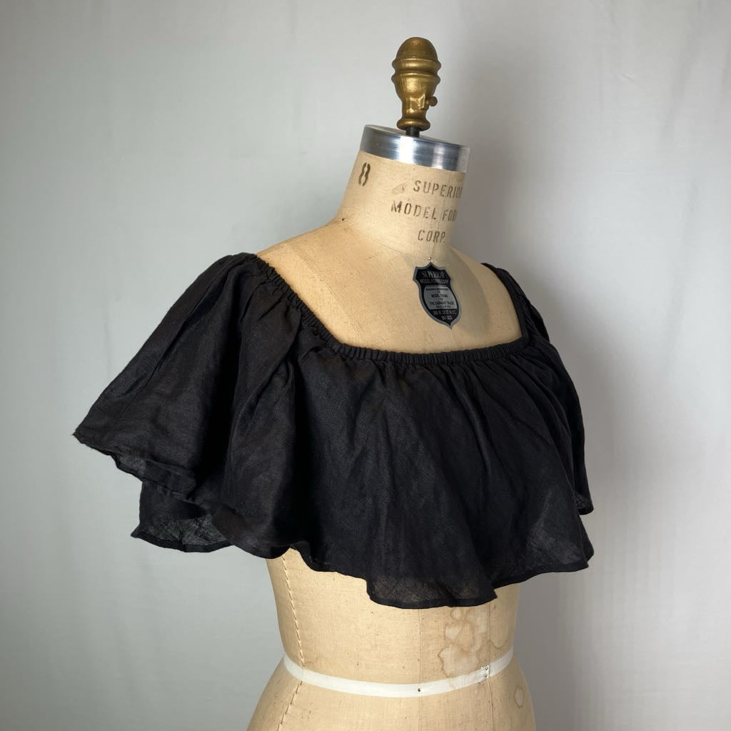 A black off-the-shoulder ruffled blouse displayed on a vintage wooden dress form mannequin against a light gray fabric backdrop.