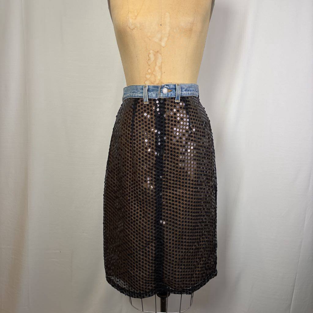 A black sequined mesh mini skirt with a light blue denim waistband, displayed on a wooden dress form against a white fabric backdrop.