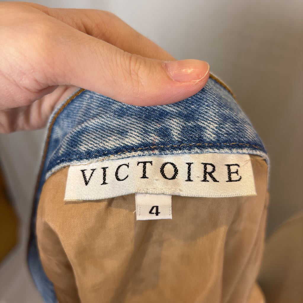 Close-up of a hand holding the collar of a garment, showing a denim blue inner lining and a sewn-in white label that reads VICTOIRE, with a size tag reading 4.