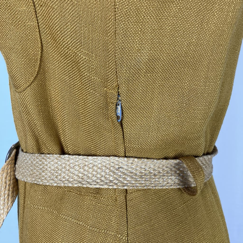 Close-up of a textured olive-brown garment with a woven beige belt, showing a central seam and a small blue-and-gold hanging tag, against a light blue background.