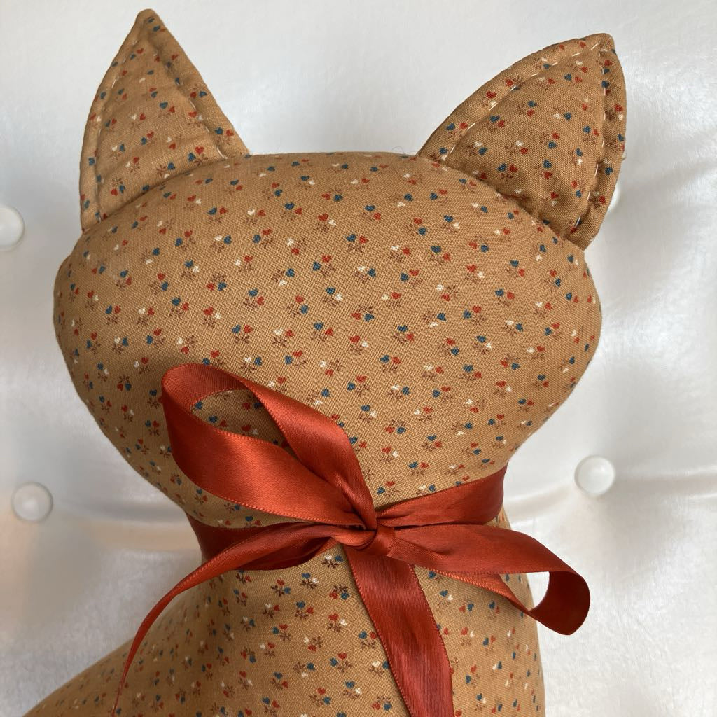 A close-up of a handmade plush cat toy with a tan floral fabric body, pointed ears, and a rust-red satin ribbon tied around its neck, set against a white tufted background.