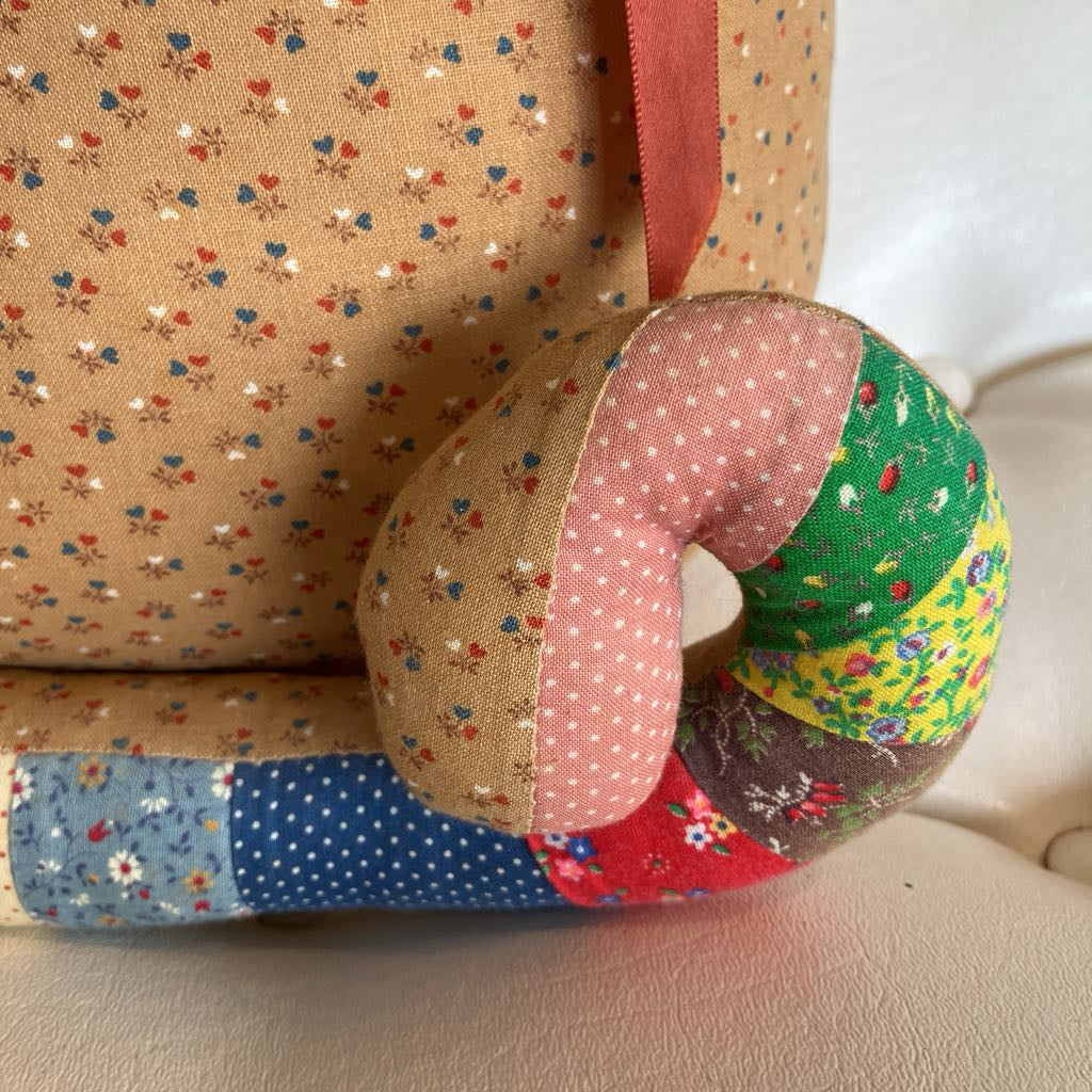 A handmade patchwork baby support pillow resting on a beige upholstered chair next to a matching floral fabric storage bag or basket.