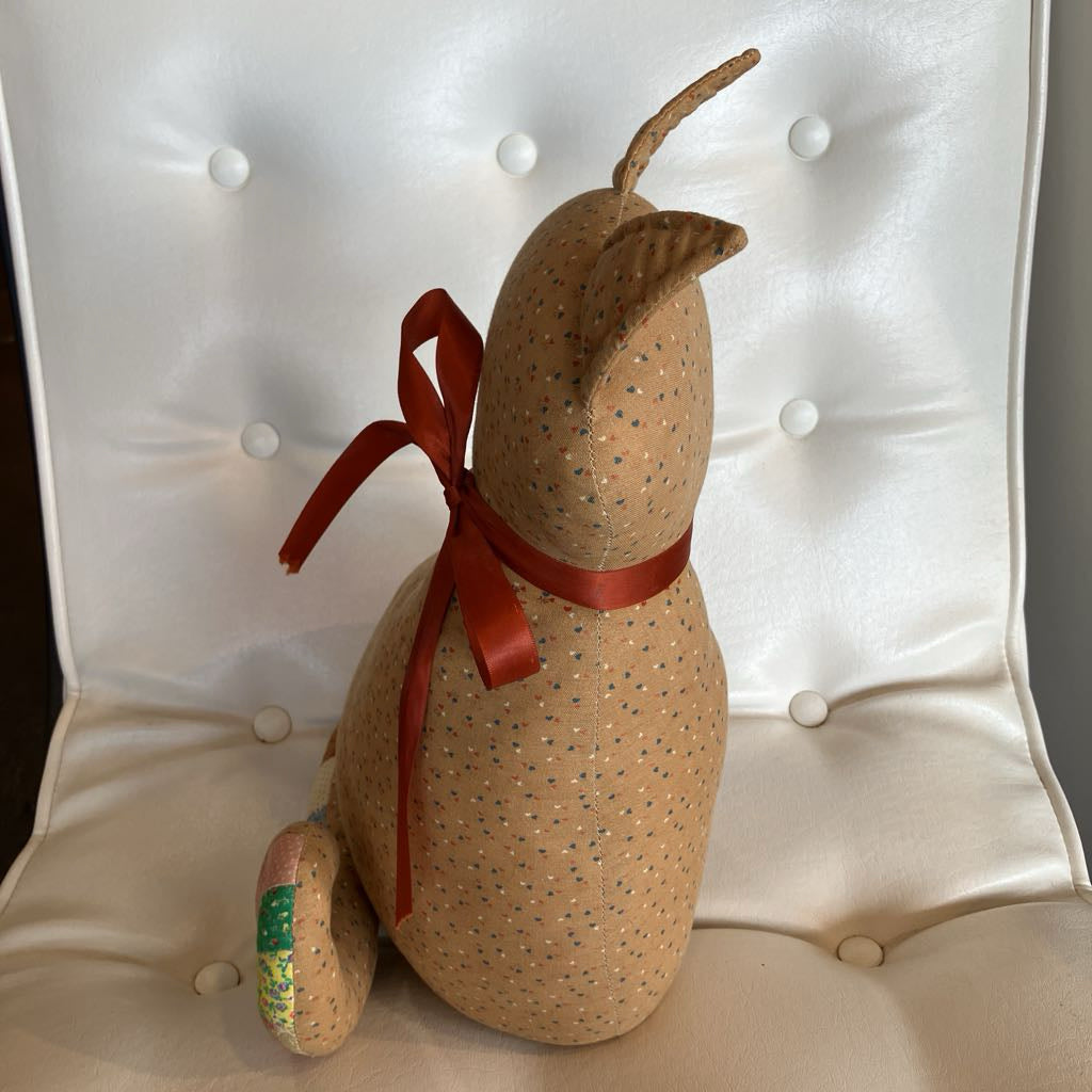 A handmade fabric dog doll with a tan body covered in tiny blue and green dots, wearing a red ribbon bow, sitting on a white tufted chair.