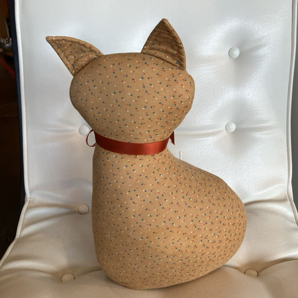 A handmade fabric cat-shaped cushion or pillow, shown from the back, featuring a tan base with a small blue and yellow polka dot pattern and a red satin collar, resting on a white tufted chair.