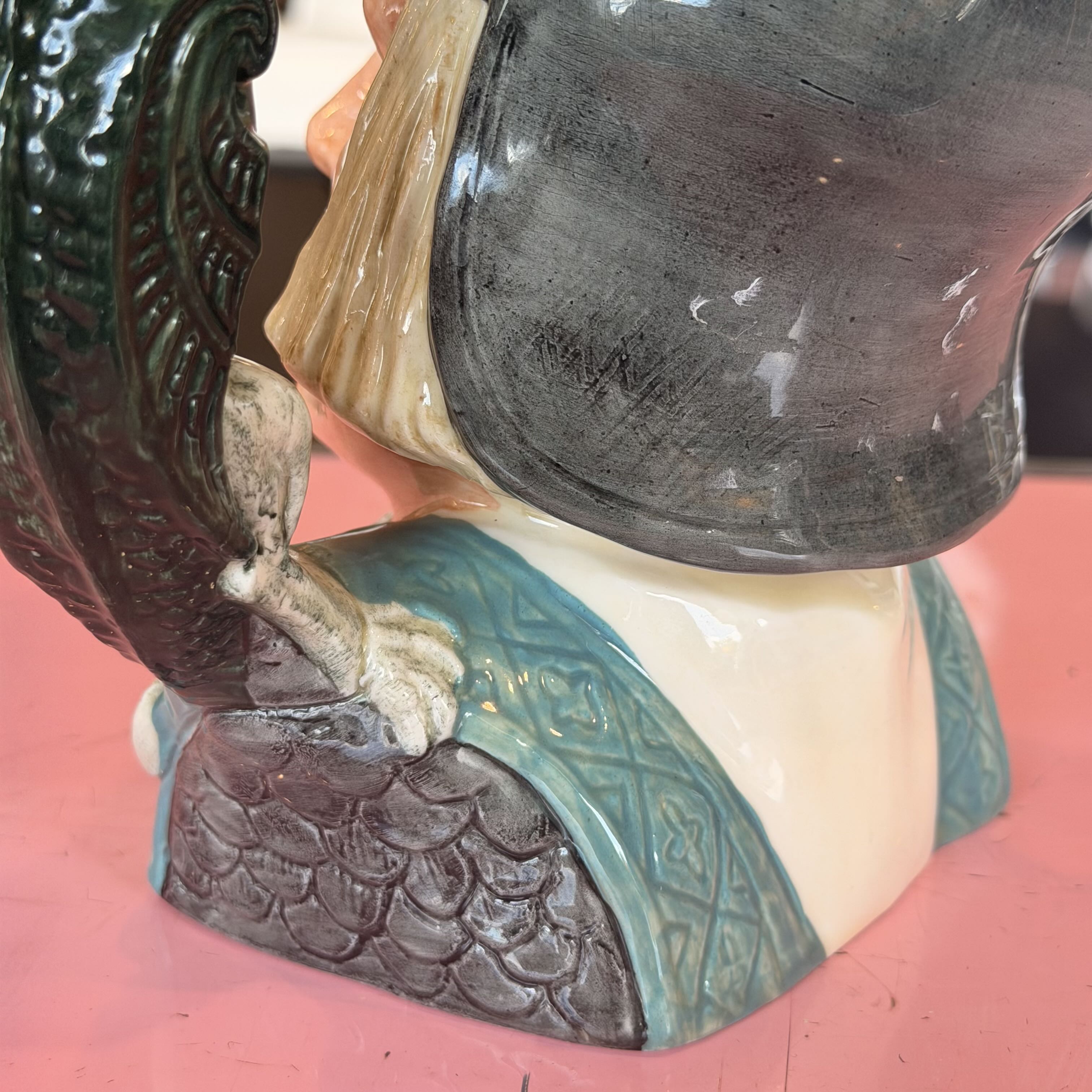 Close-up of a ceramic figurine showing part of a rooster with a glossy dark green beak and tail, a cream-colored body, and a teal and dark gray patterned garment, set against a pink background.