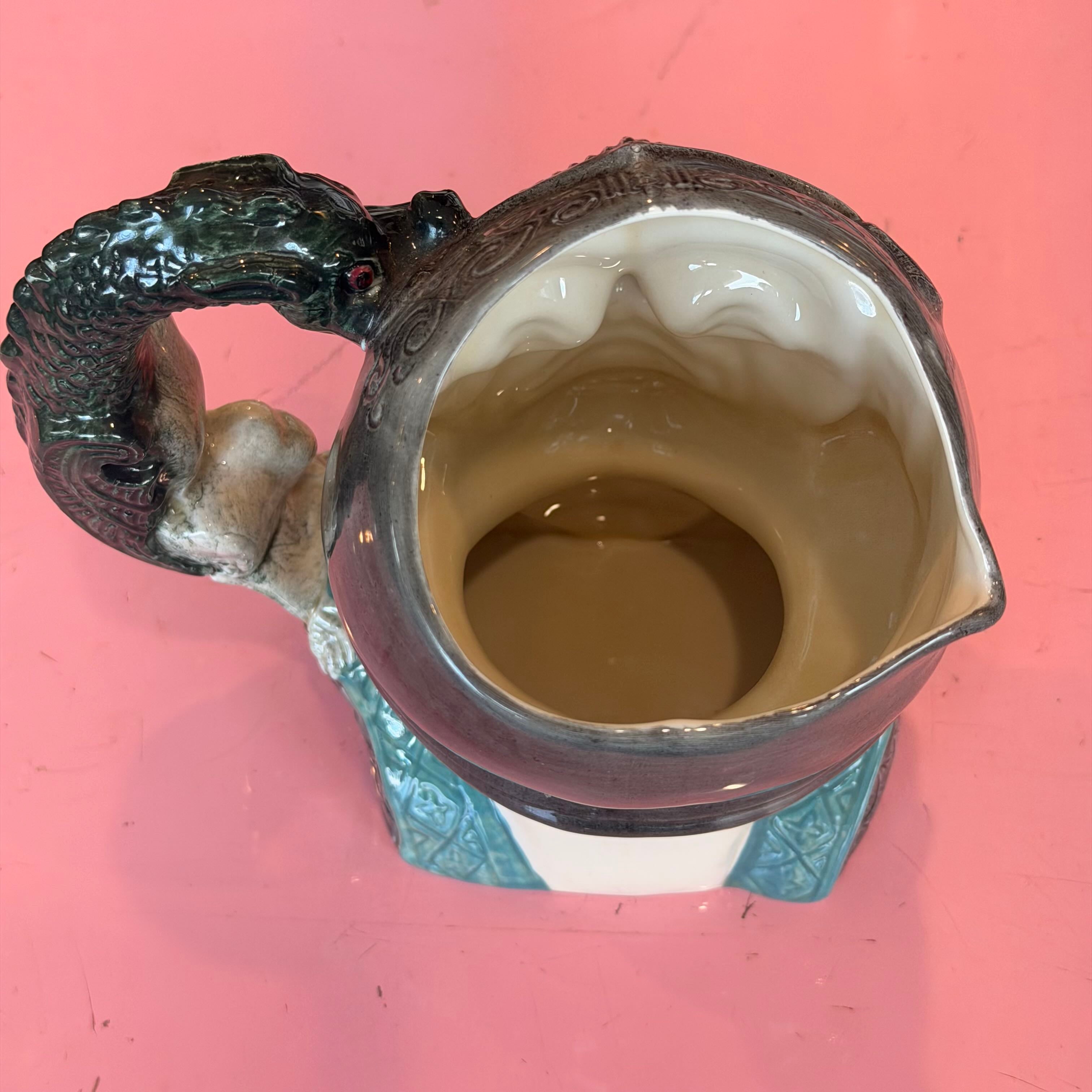 A ceramic pitcher with a sculpted handle shaped like a dragon, featuring a dark gray exterior, a beige interior, and a turquoise blue base, shown on a pink background.