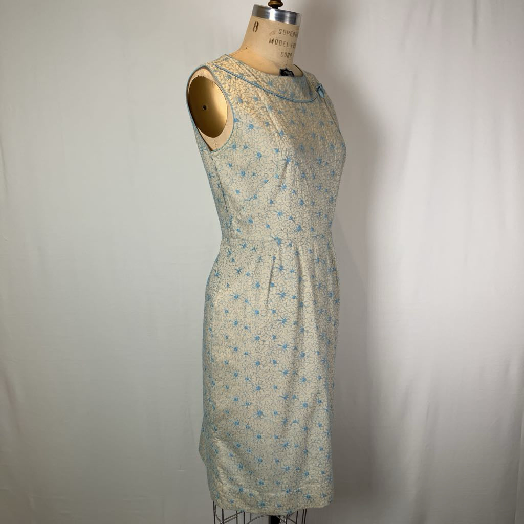 A sleeveless knee-length dress with a small blue floral pattern on a light beige or off-white base, displayed on a vintage dress form mannequin against a white fabric backdrop.