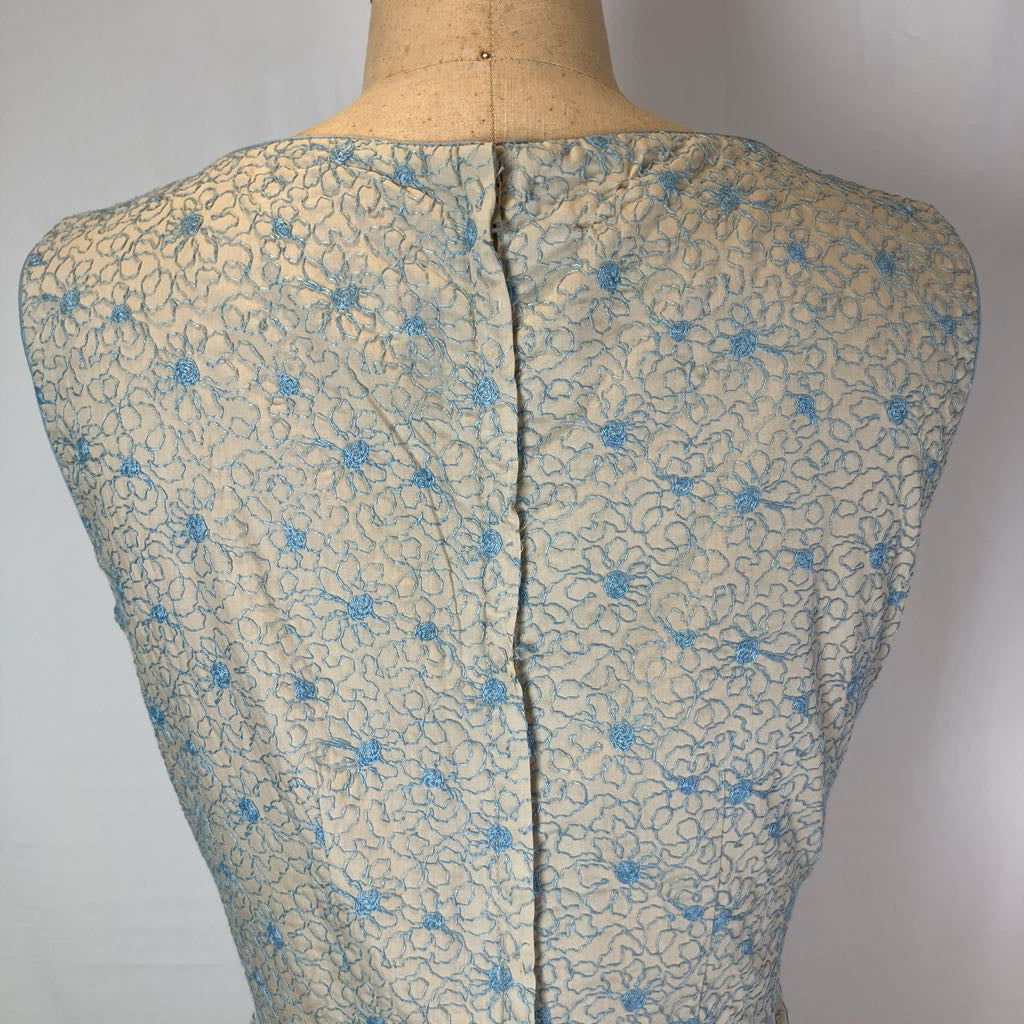 The back view of a vintage sleeveless dress or blouse displayed on a fabric mannequin torso, featuring a light beige base with a blue floral and vine pattern and a central seam.