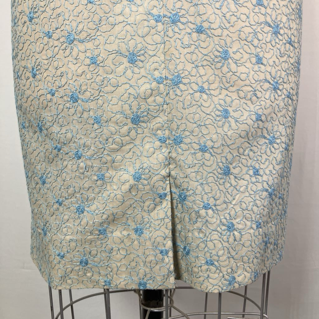 A close-up of a skirt or shorts garment displayed on a dress form, featuring a beige base with an allover light blue floral and scroll pattern.