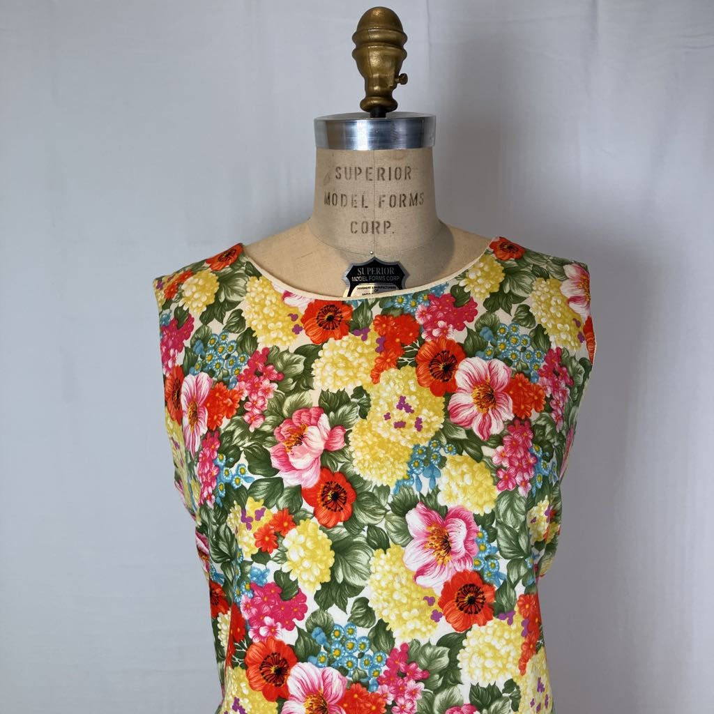 A sleeveless vintage-style top or dress on a dress form, featuring a dense multicolor floral print with yellow, red, pink, blue, and green tones on a light background.