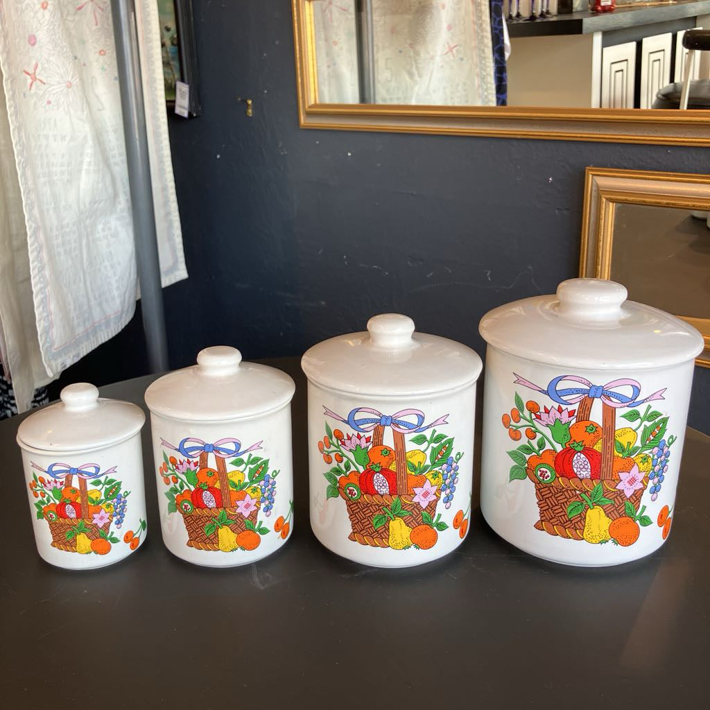 Four white ceramic canisters with lids, arranged by size on a dark tabletop. Each canister features a colorful basket illustration filled with fruits and flowers, tied with a blue ribbon motif.