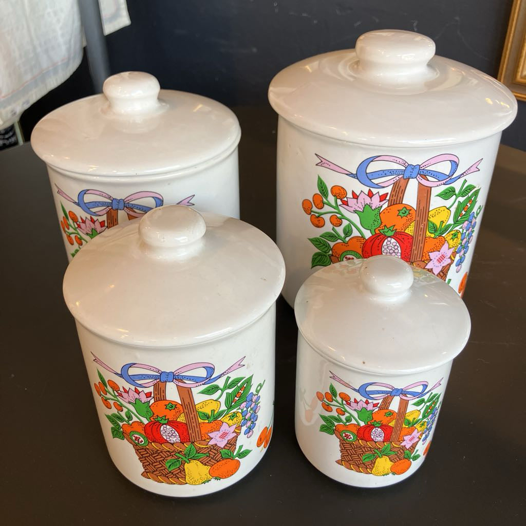 Three white ceramic canisters with matching lids, decorated with colorful baskets of fruit and flowers and purple bows, arranged on a dark tabletop.