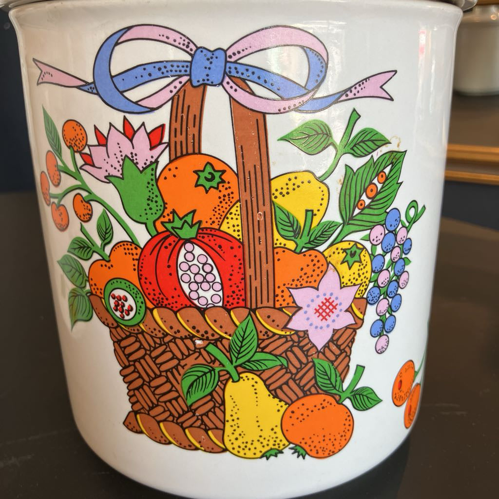A white ceramic canister or cup featuring a colorful retro illustration of a fruit and vegetable basket with a pink and blue bow, set against a dark background.