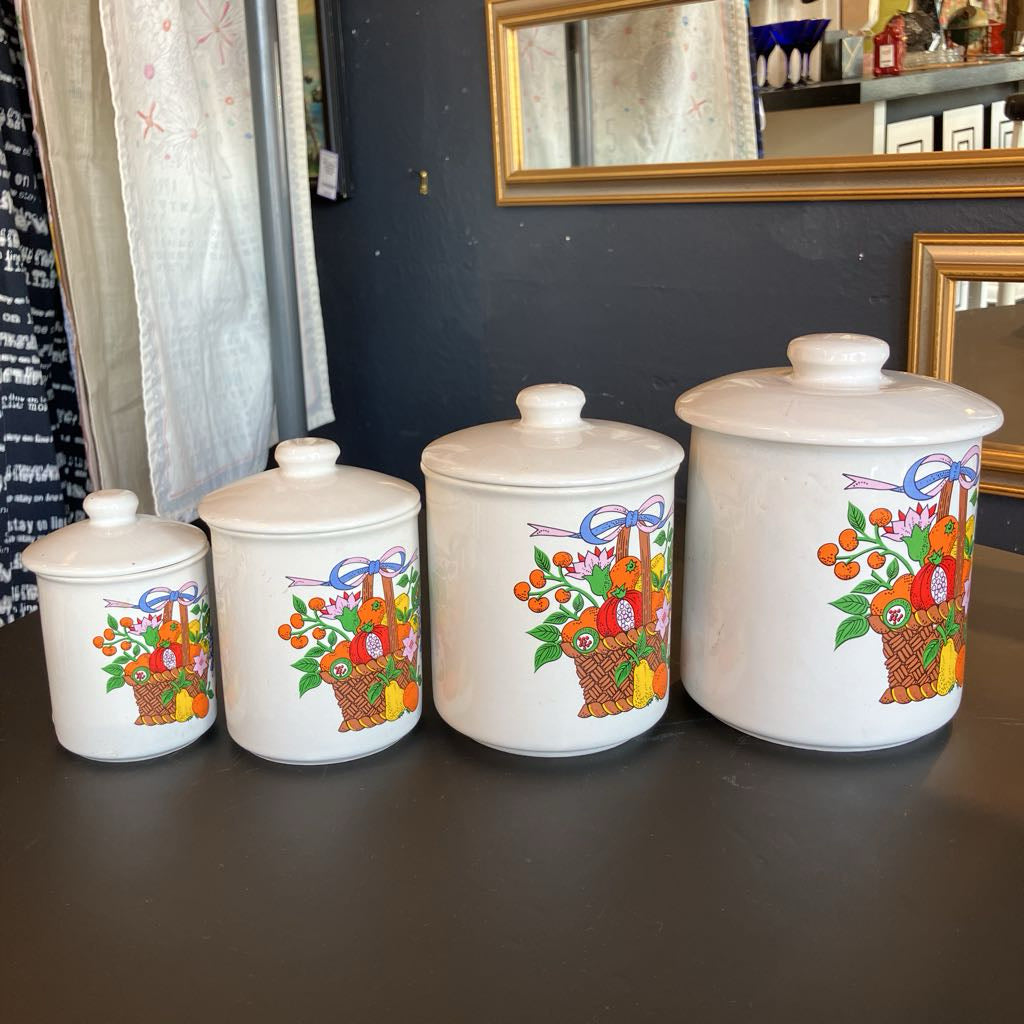 Three white ceramic canisters with lids, arranged by size on a dark tabletop. Each canister features a colorful basket of fruit and vegetables printed on the front, with purple bows above the baskets.