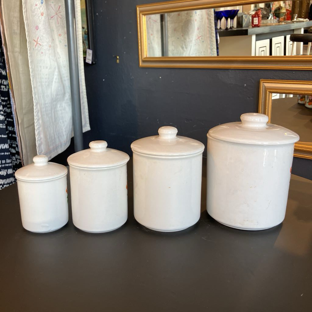Four white ceramic canisters of varying sizes with matching lids, arranged in a row on a dark tabletop.