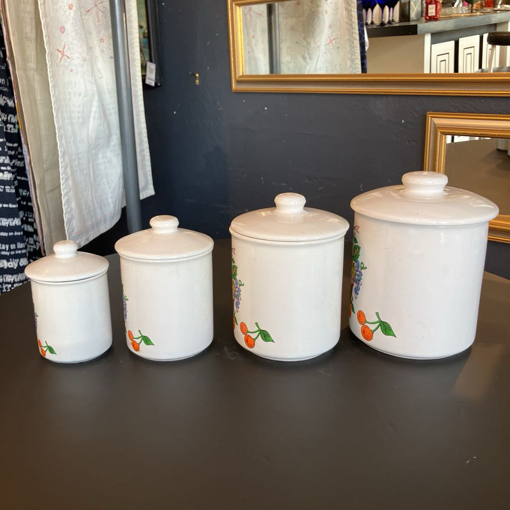 Four white ceramic canisters with matching lids, arranged by size on a dark tabletop. Each canister features a colorful fruit illustration, including orange, purple, and green.