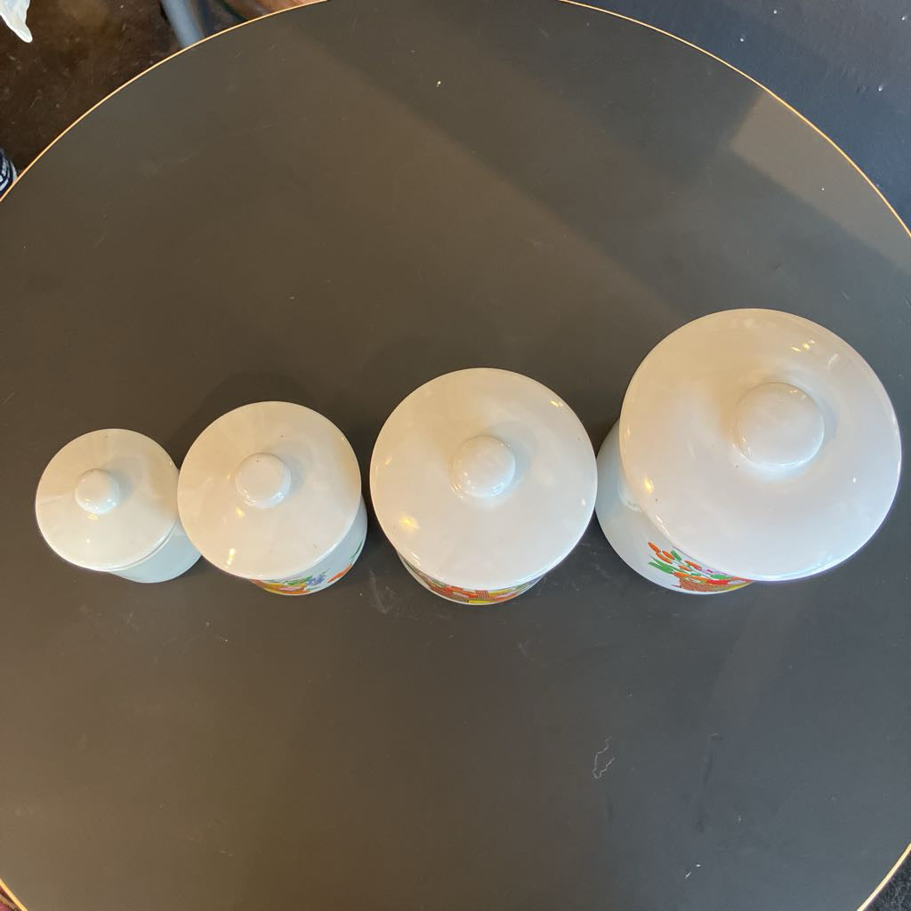 Four white ceramic lidded canisters in a graduated size set, arranged in a row on a dark reflective surface.