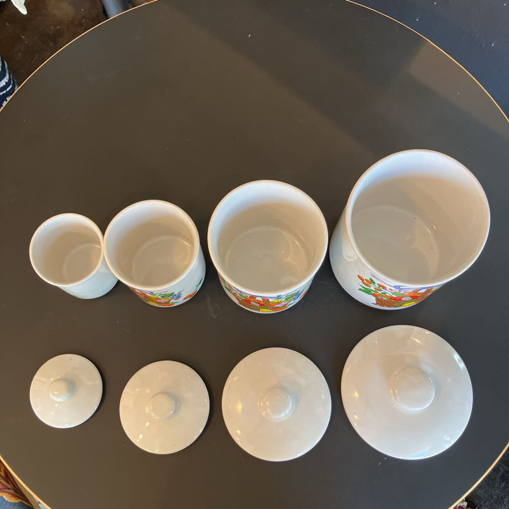 A set of six white ceramic tumblers with matching lids, arranged by size on a dark round table. The tumblers feature a colorful printed design near the base.