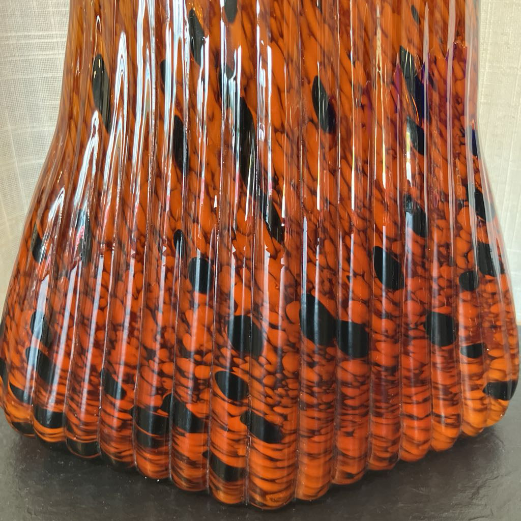 A close-up of a glossy, fluted glass vase or container with a vibrant orange and black marbled pattern, set against a neutral beige background.