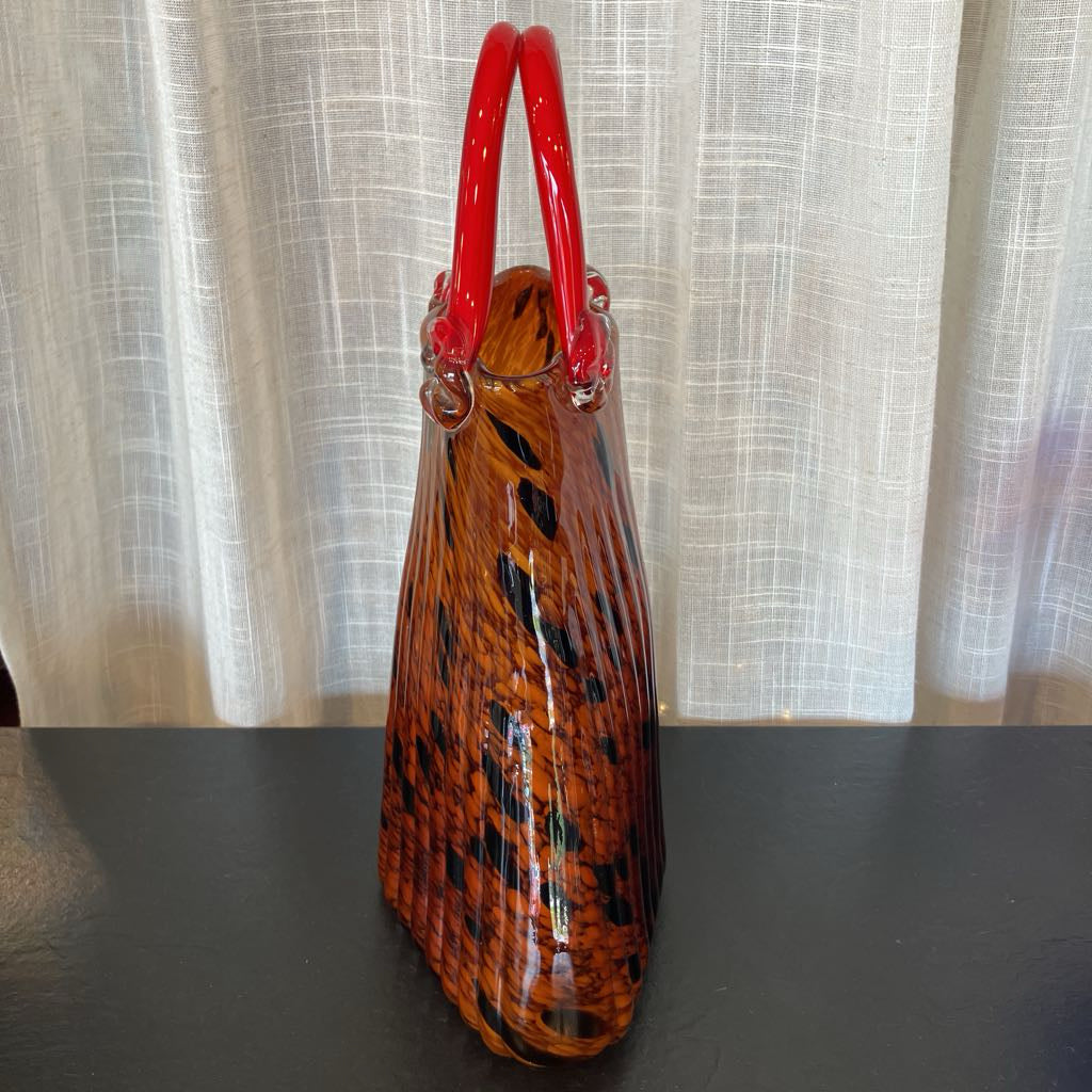 A hand-blown glass handbag with a glossy orange and black patterned body and bright red double top handles, standing on a dark surface in front of a light curtain backdrop.