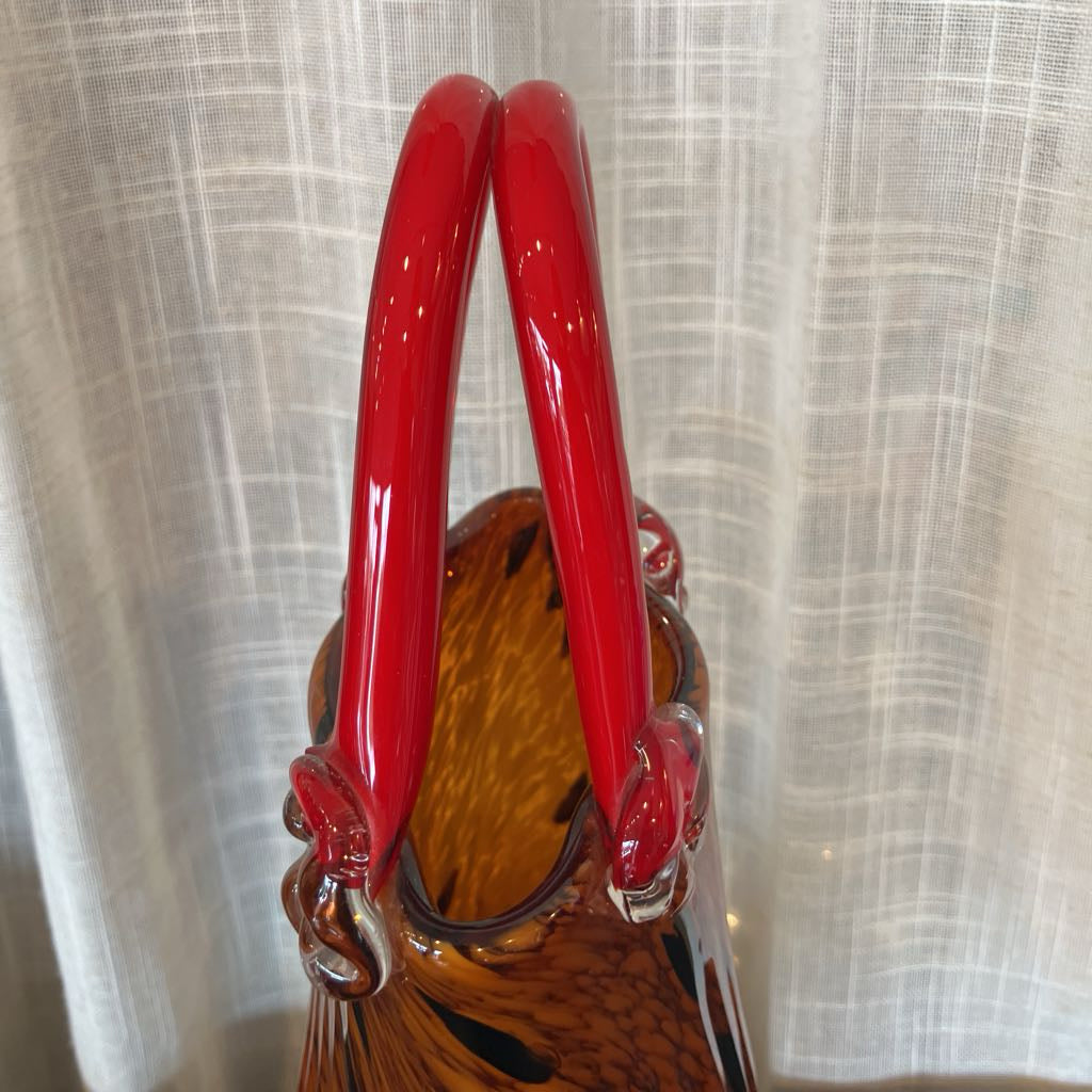 A close-up of a hand-blown glass bag or vase with glossy red handles and a smoky amber-brown glass body, set against a light beige curtain background.