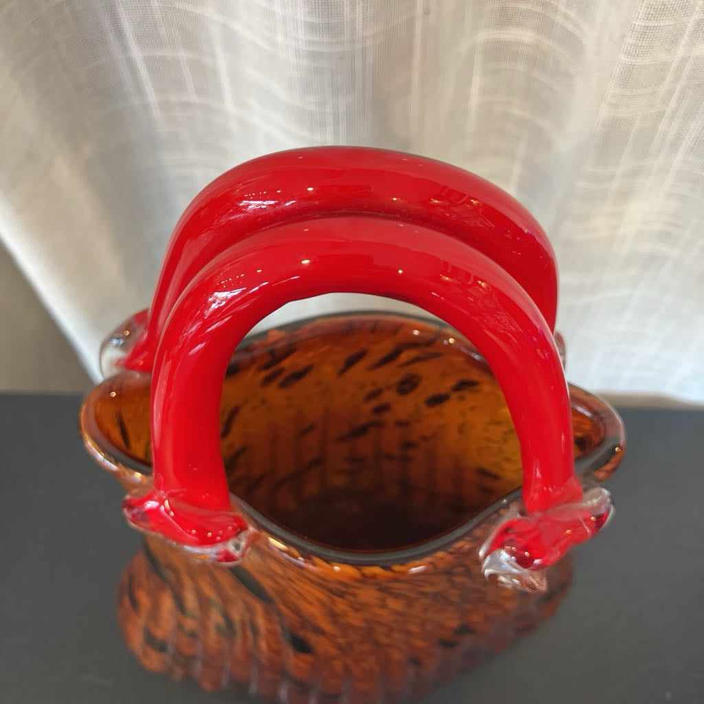 A small decorative basket with glossy red double handles and a mottled amber-brown glass body, photographed against a light curtain background.