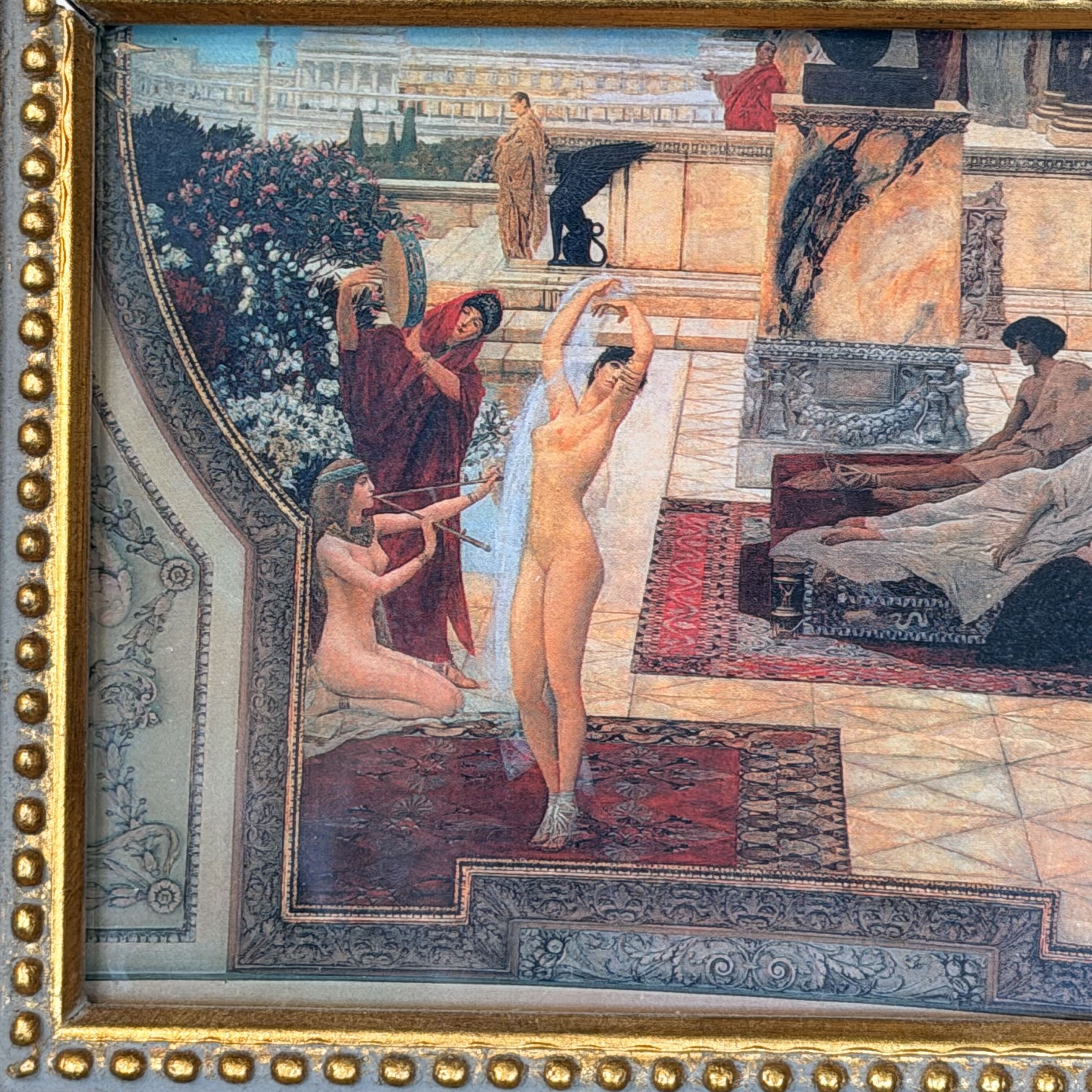 A framed classical painting depicting several nude figures in an ornate interior scene with red rugs, floral arrangements, and a gold beaded frame.