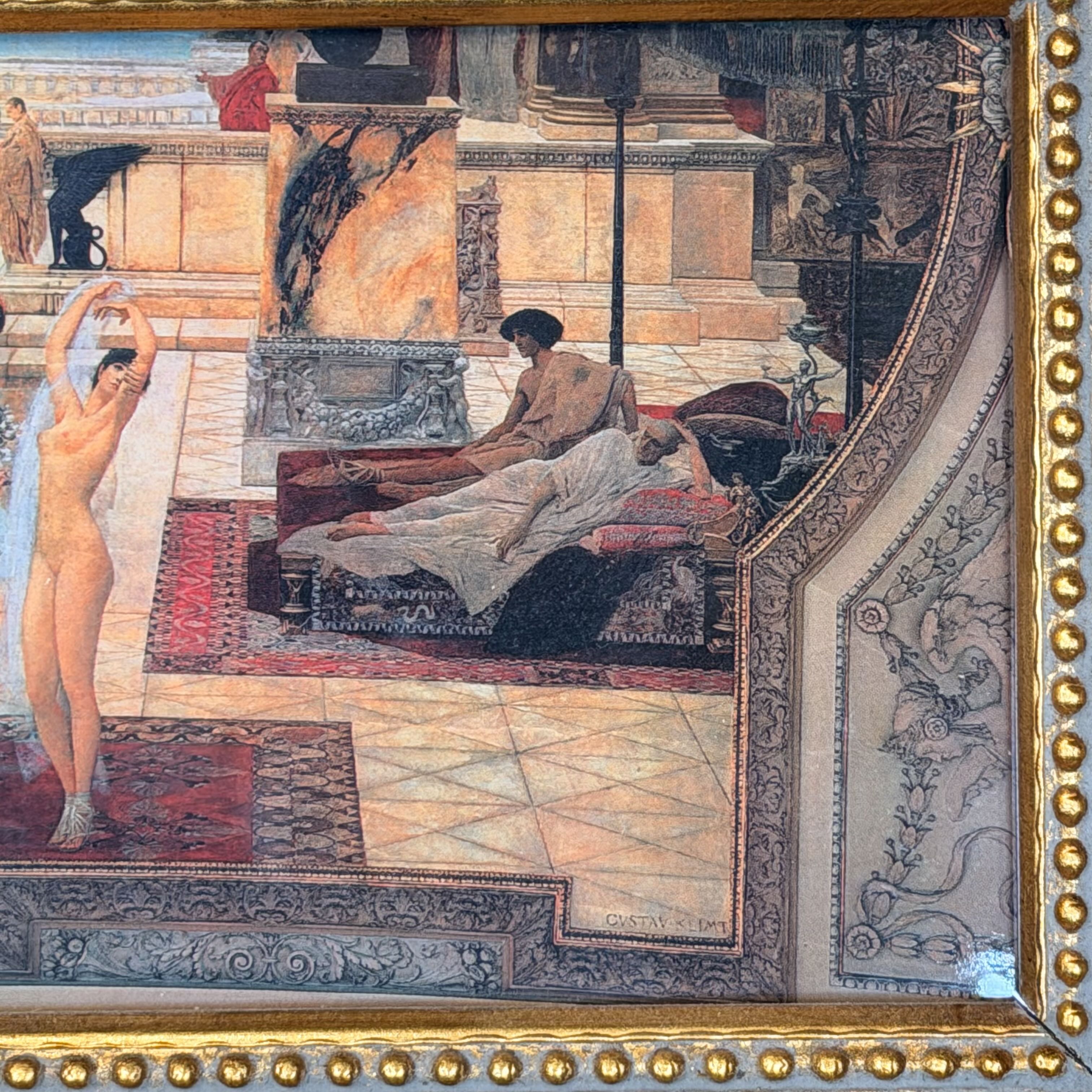 A framed classical painting depicting a luxurious interior scene with several nude and semi-nude figures in an ornate room with patterned rugs, marble-like walls, and a gold beaded frame bordering the artwork.