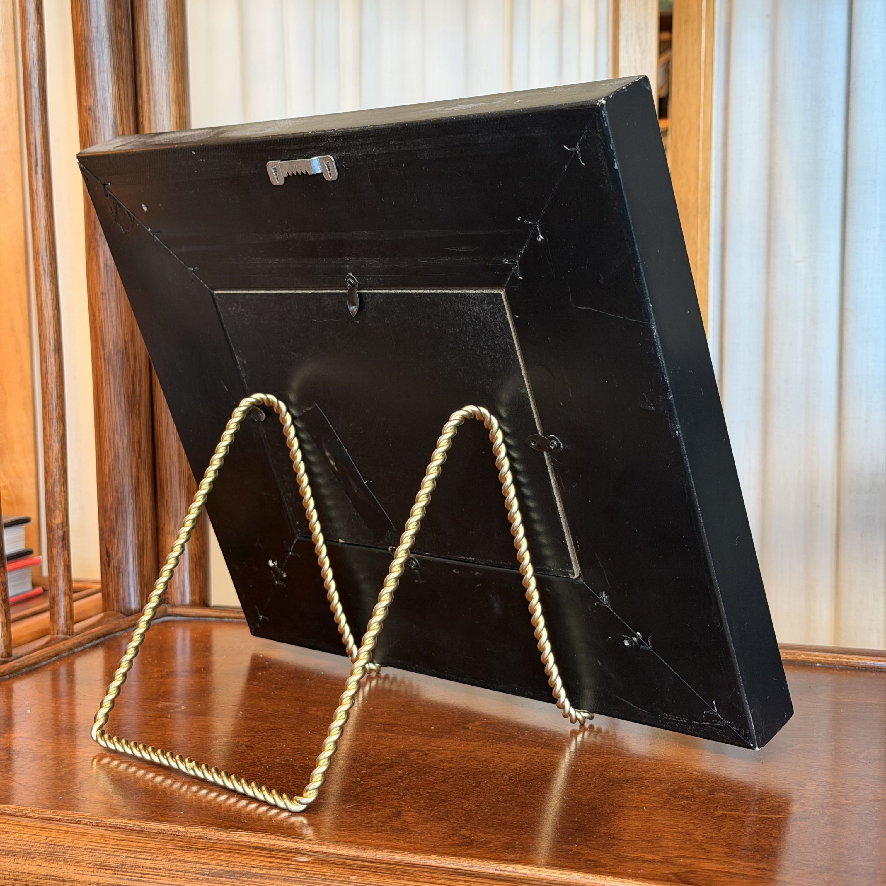 A black rectangular picture frame shown from the back, propped open on a twisted gold metal easel stand on a wooden table, with a home interior background.