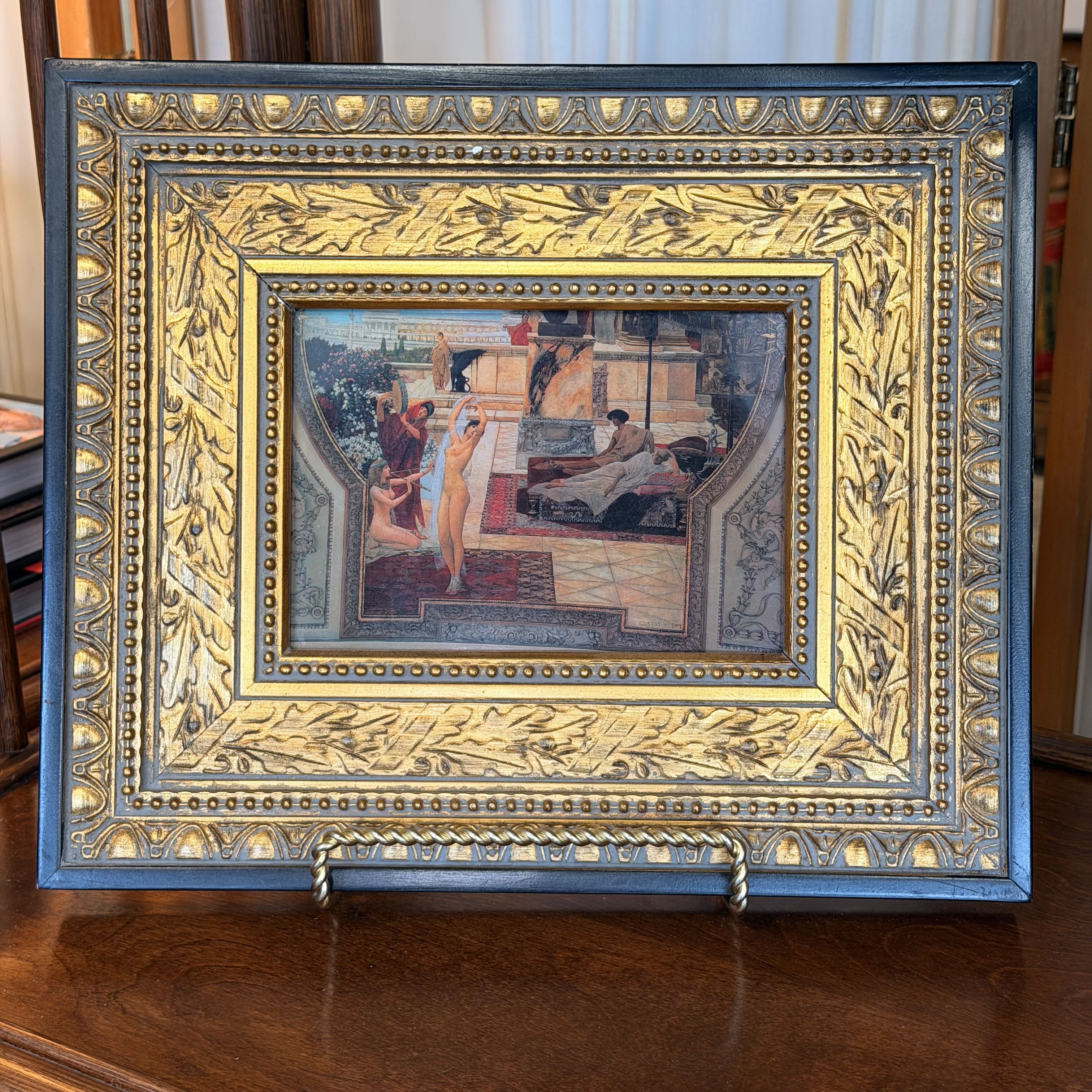 An ornate framed art piece featuring a central miniature painting of figures in an interior scene, surrounded by a highly decorative gilded and blue frame with embossed floral and geometric patterns, displayed on a wooden stand on a wooden table.