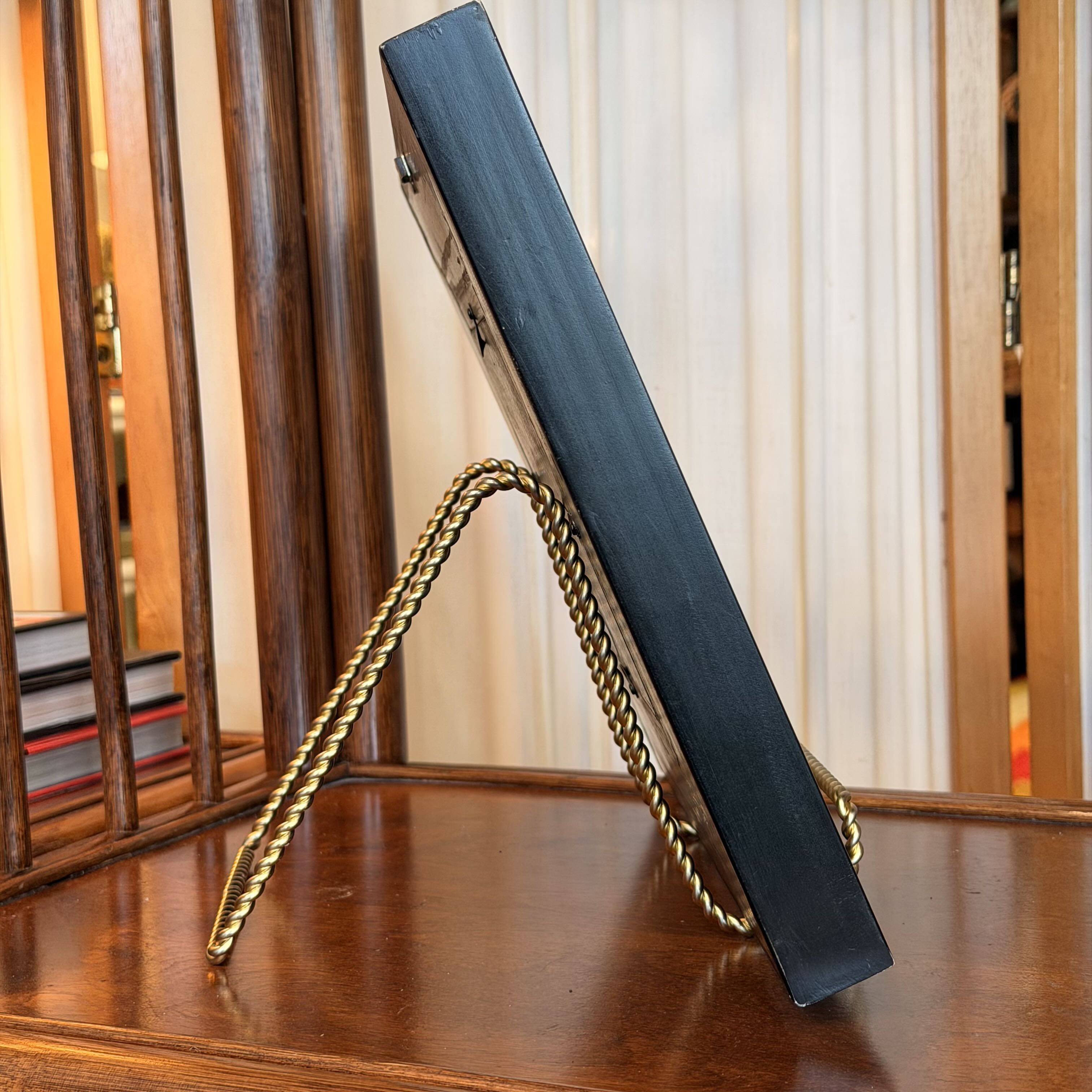 A black rectangular picture frame or book-like object propped upright on a twisted gold metal stand, placed on a polished wooden table with a warm indoor background.