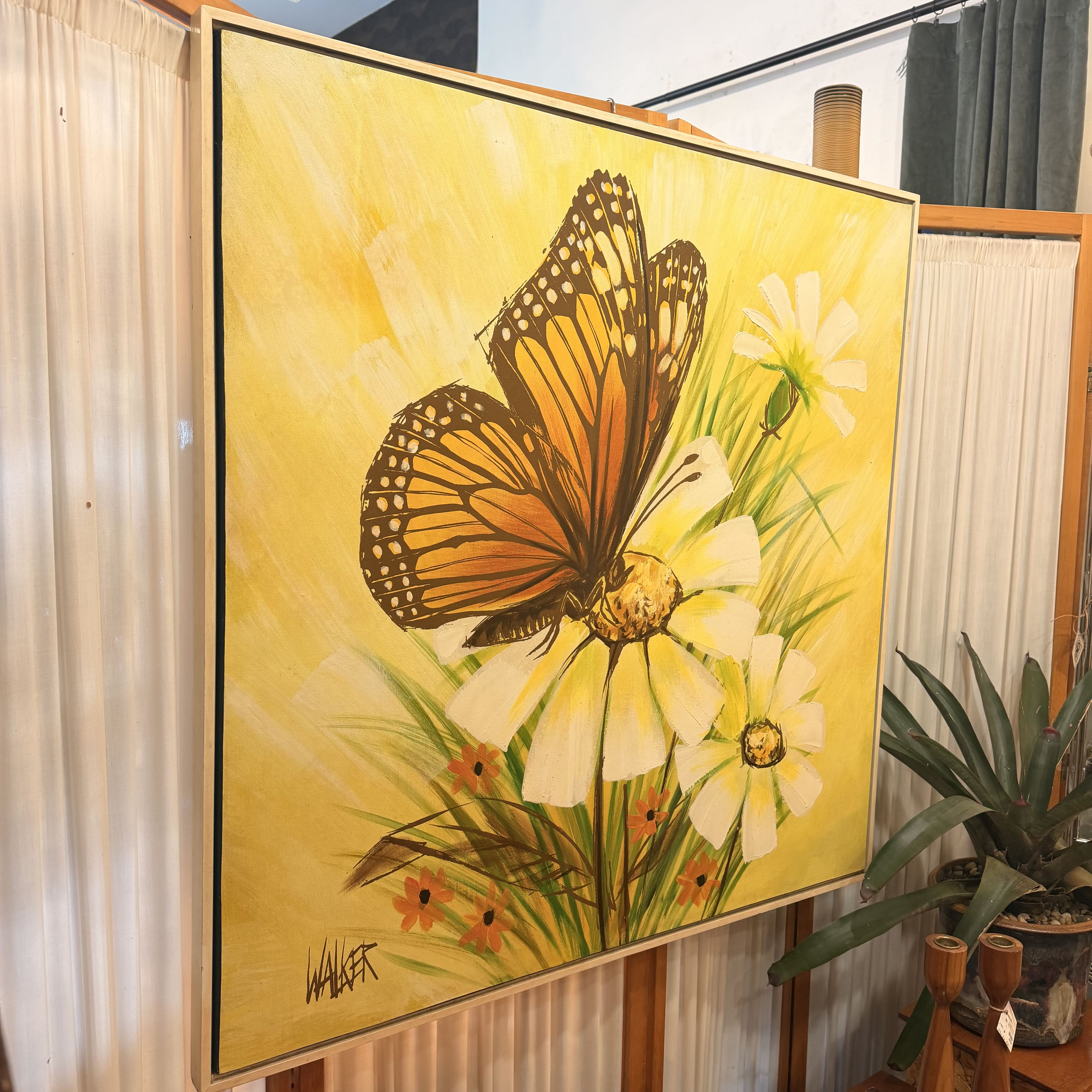 A large framed canvas painting featuring an orange and black butterfly perched on white flowers against a bright yellow background, displayed indoors on a wooden stand with curtains and a potted plant nearby.