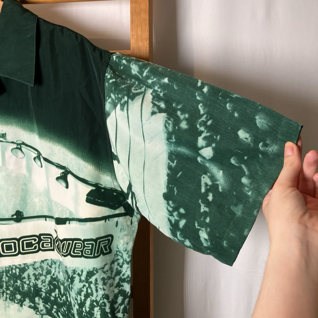 A close-up of a green short-sleeve button-up shirt with a tie-dye or watercolor-style print, held by a hand against a light wall.