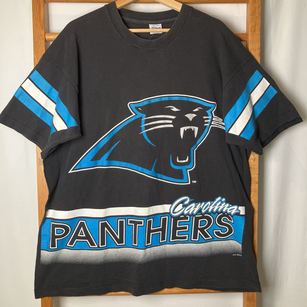 A black Carolina Panthers graphic t-shirt hanging on a wooden hanger against a white curtain backdrop. The shirt features the team's panther logo in blue and white, with 'Carolina PANTHERS' text across the bottom.
