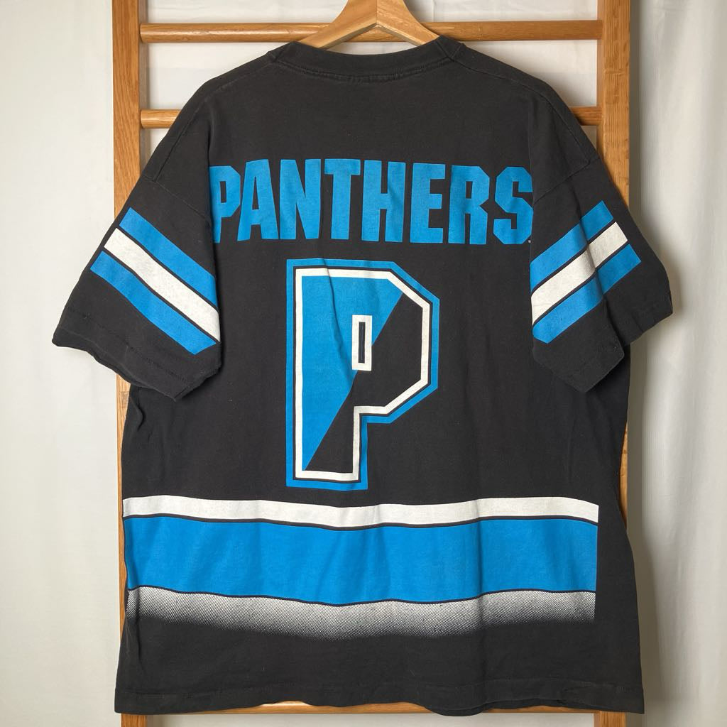 A black short-sleeve t-shirt with a Panthers theme, featuring a large blue and white letter 'P' and the word 'PANTHERS' on the back, hanging on a wooden hanger against a light wall.