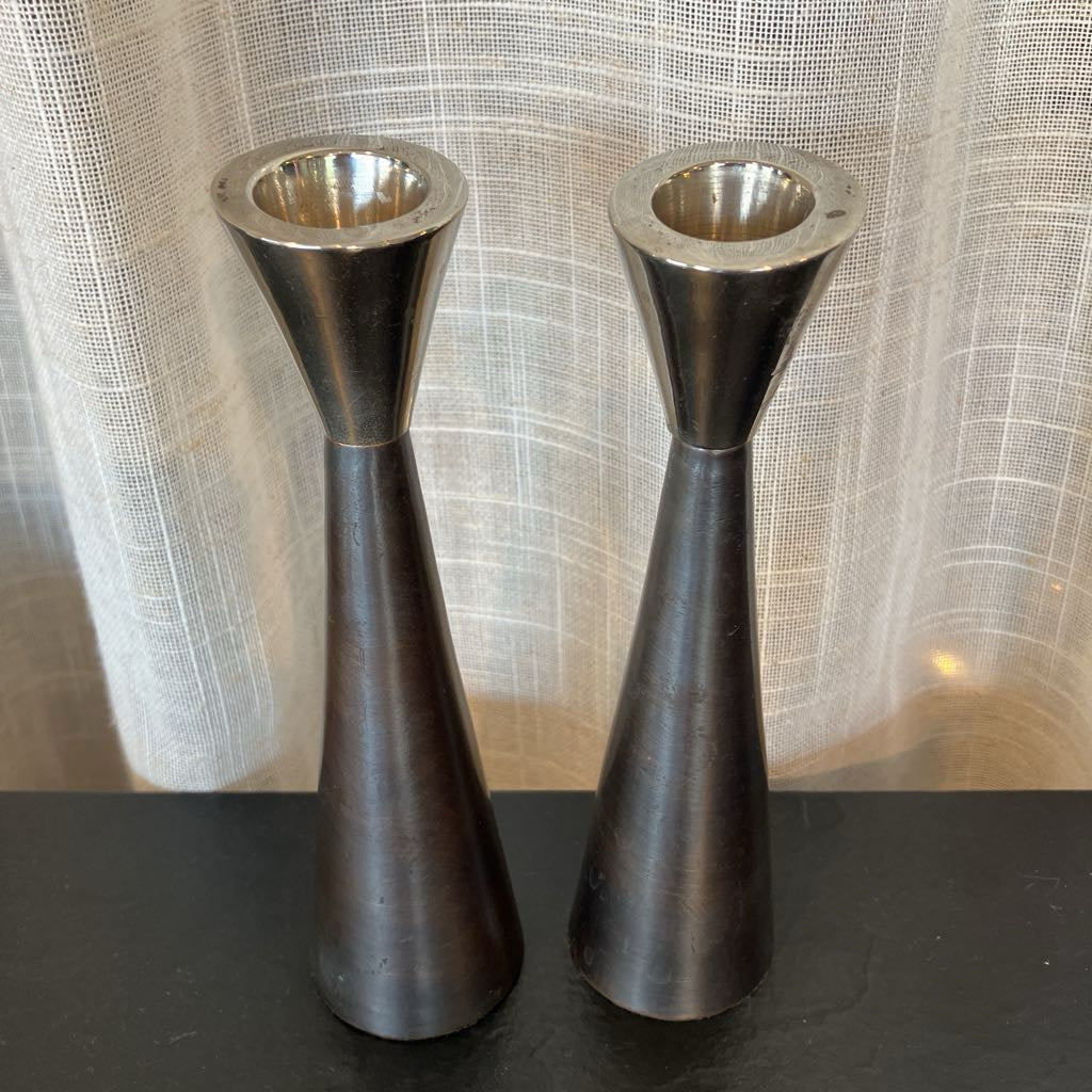 A pair of vintage dark metal candlestick holders with a flared top and tapered base, standing on a dark surface in front of a white textured curtain.