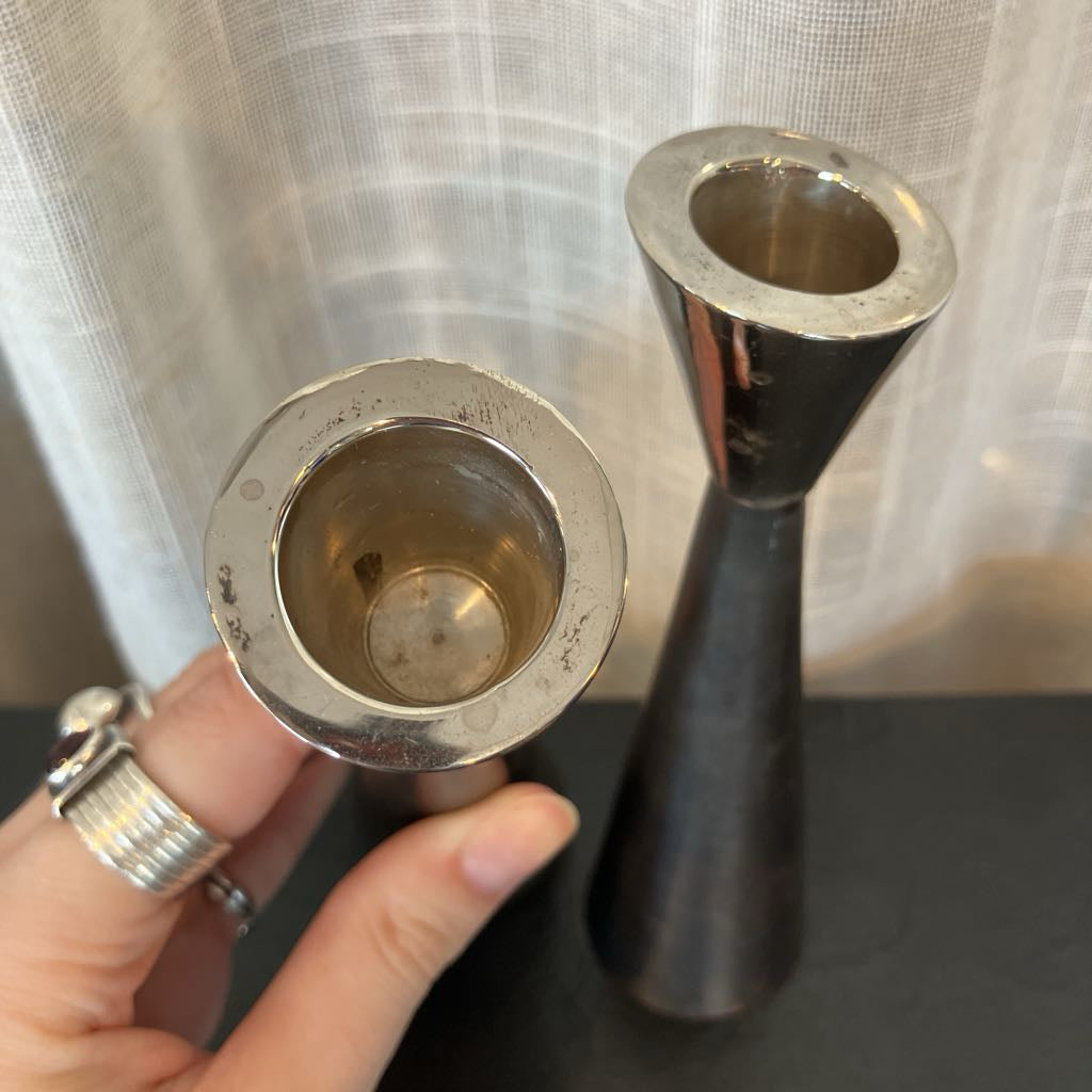 A close-up of a hand holding a small silver-toned metal cup or tumbler, with a taller dark metal vase or candle holder standing behind it against a sheer curtain backdrop.