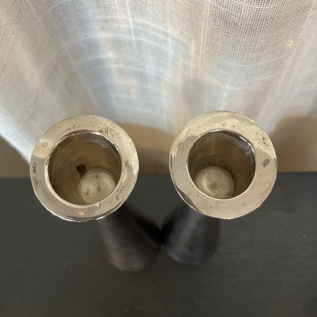 Two silver metal candlestick holders or candle bases with circular cutouts, shown from above on a dark surface with a sheer curtain in the background.