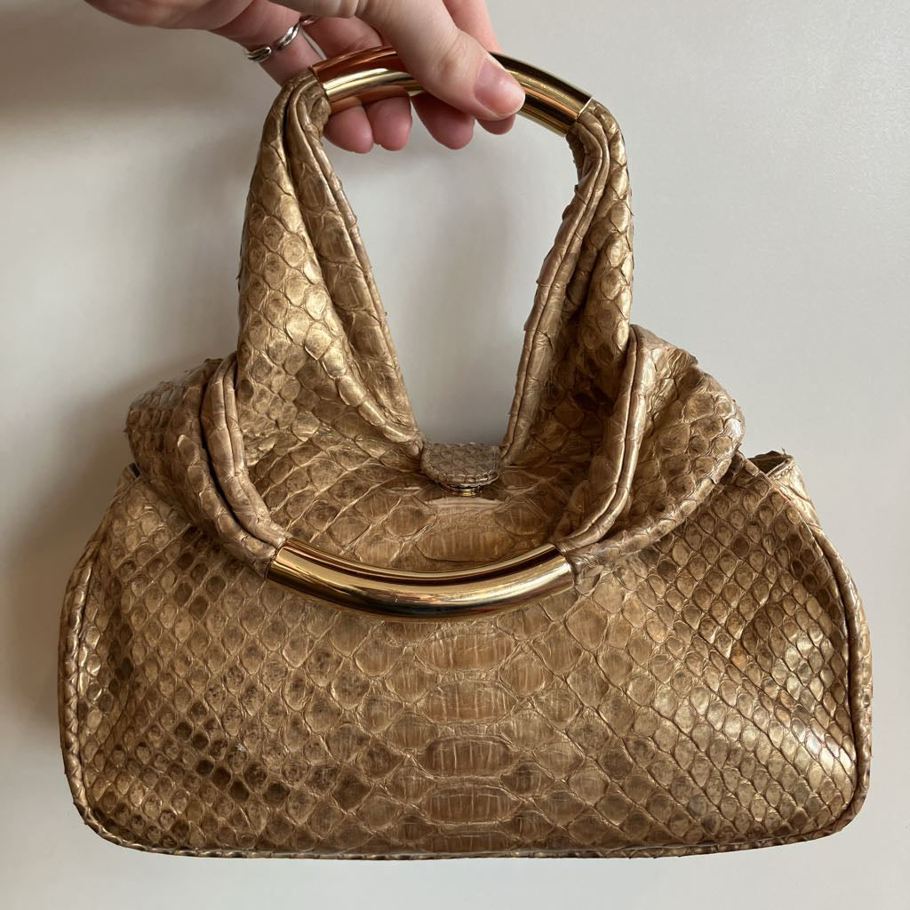 A gold-toned snakeskin handbag with a curved metal top handle, held by a hand against a plain off-white background.