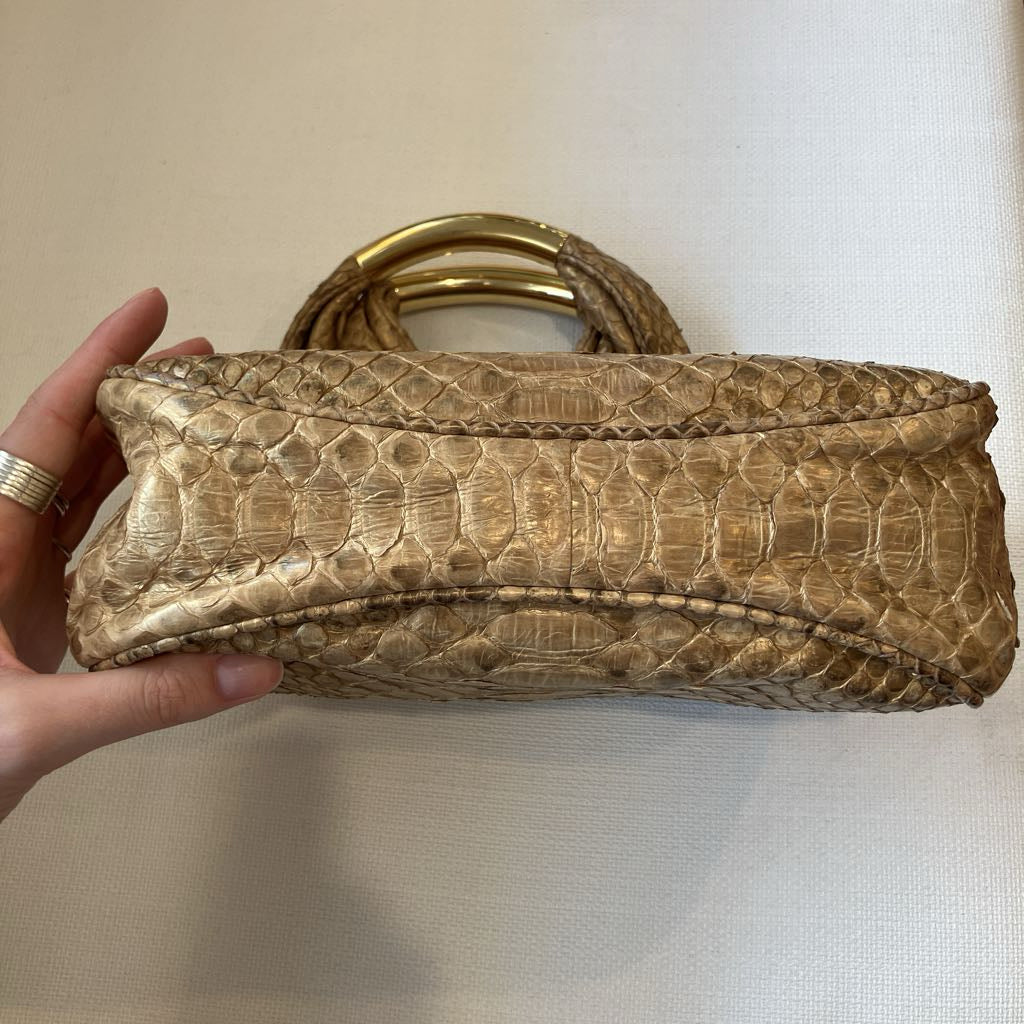 A hand holding a small structured handbag made of textured reptile skin in a light tan color, featuring a gold-tone metal ring handle, set against a neutral off-white background.