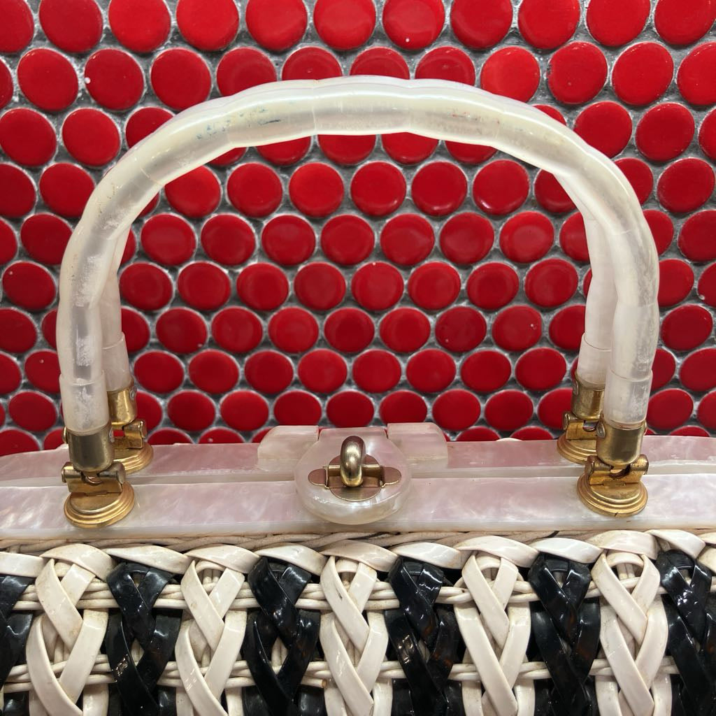 A close-up of a handbag featuring a translucent white resin handle, gold-tone hardware, and a black and white woven body, set against a red circular tile background.