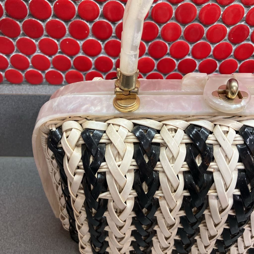 A close-up of a woven handbag with cream and black braided leather straps, a translucent pink shell top, and gold-tone hardware, set against a red circular tile background.